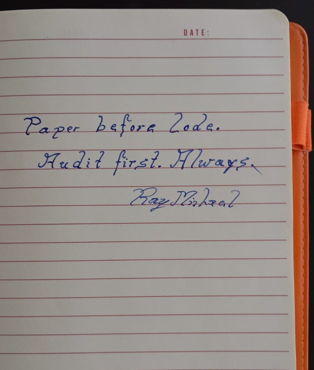 An open notebook with 'Paper before code. Audit first. Always.' written in blue ink, an orange pen lying across the page